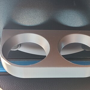 May include: A gray plastic cup holder with two circular openings. The holder is mounted on a black surface.