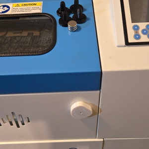 May include: A white laser cutter with a blue top and a black and white control panel. The control panel has a screen and buttons. The laser cutter has a safety warning sign that says "Caution: Wear eye protection during operation!"