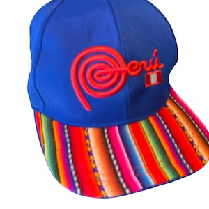 May include: A blue baseball cap featuring the word "Perú" embroidered in red thread with a spiral design and a small Peruvian flag icon. The brim displays a vibrant, multi-colored striped pattern in shades of red, orange, yellow, green, and purple.