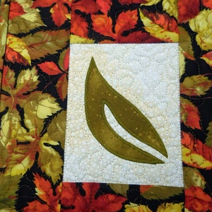 以下が含まれることがあります： 秋の葉のデザインが施されたキルティング生地のプレースマット。プレースマットの中央には大きな緑の葉があり、その周りを赤、オレンジ、黄色の小さな葉が囲んでいます。