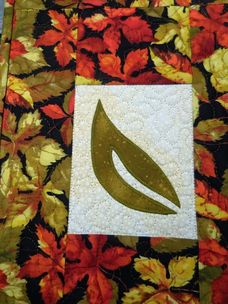 以下が含まれることがあります： 秋の葉のデザインが施されたキルティング生地のプレースマット。プレースマットの中央には大きな緑の葉があり、その周りを赤、オレンジ、黄色の小さな葉が囲んでいます。