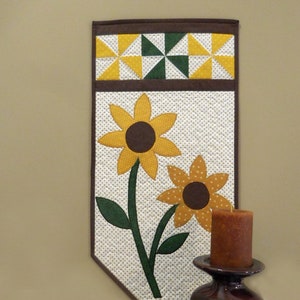 May include: A quilted wall hanging with a brown border and a white background with a pattern of small yellow dots. The centre of the hanging features two yellow sunflowers with brown centres and green stems and leaves.