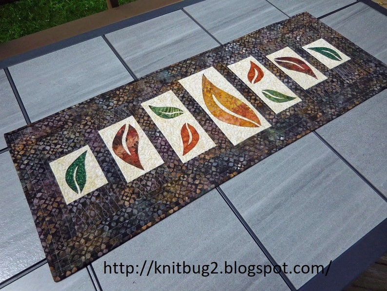 以下が含まれることがあります： 濃い茶色の背景に、緑、オレンジ、茶色の葉の模様が施されたテーブルランナー。葉は白で縁取られ、ランナーの中央に一列に並んでいます。ランナーは布製で、フリンジの縁取りが施されています。ランナーの下部には「http://knitbug2.blogspot.com/」というテキストが印刷されています。