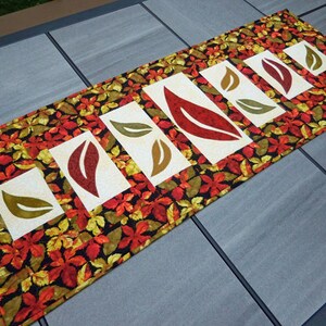以下が含まれることがあります： 黒の背景に赤、黄、緑の葉の模様が施されたテーブルランナー。ランナーには、赤、黄、緑の葉が切り抜かれた白い正方形が5つあります。