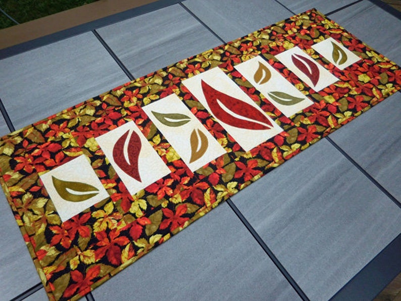 以下が含まれることがあります： 黒の背景に赤、黄、緑の葉の模様が施されたテーブルランナー。ランナーには、赤、黄、緑の葉が切り抜かれた白い正方形が5つあります。