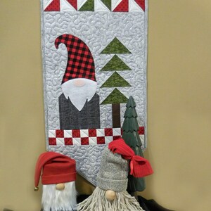 May include: Two gnomes, one with a red hat and the other with a gray hat, stand in front of a quilted wall hanging. The wall hanging features a red, white, and green patchwork design with a gnome and trees.