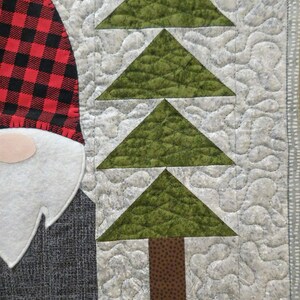 May include: A quilted fabric wall hanging with a red and black plaid gnome and a green tree with a brown trunk. The background is a light gray with a quilted pattern.