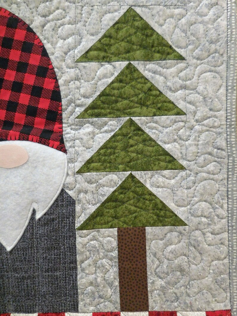 May include: A quilted fabric wall hanging with a red and black plaid gnome and a green tree with a brown trunk. The background is a light gray with a quilted pattern.