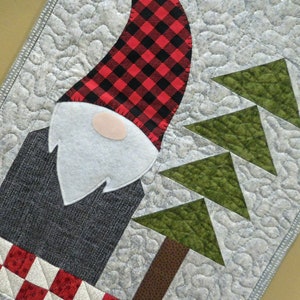 May include: A quilted table runner with a gnome design. The gnome is wearing a red and black plaid hat and a white beard. The background is a light gray with green trees.