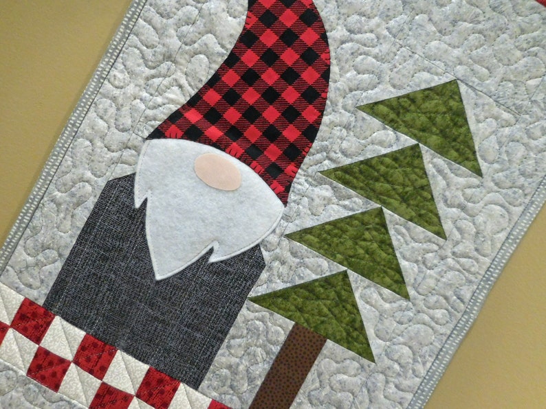 May include: A quilted table runner with a gnome design. The gnome is wearing a red and black plaid hat and a white beard. The background is a light gray with green trees.