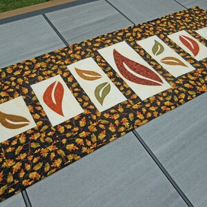 以下が含まれることがあります： 黒地に小さなオレンジと緑の葉の模様が施された長いテーブルランナー。ランナーには、オレンジ、ブラウン、グリーンの大きな葉がスタイリッシュに描かれた白いパネルが5つあります。