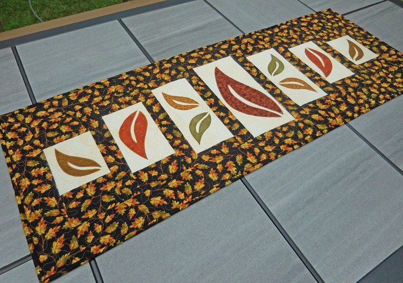 以下が含まれることがあります： 黒地に小さなオレンジと緑の葉の模様が施された長いテーブルランナー。ランナーには、オレンジ、ブラウン、グリーンの大きな葉がスタイリッシュに描かれた白いパネルが5つあります。