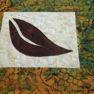 以下が含まれることがあります： 緑と金の色の花柄の白い背景に、茶色の葉の形をした布のアップリケが施されています