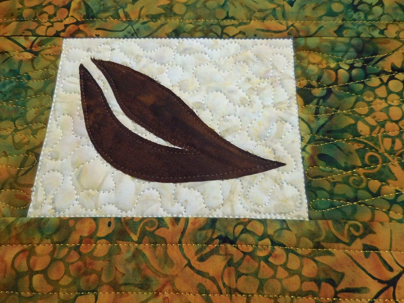 以下が含まれることがあります： 緑と金の色の花柄の白い背景に、茶色の葉の形をした布のアップリケが施されています