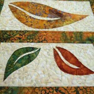 以下が含まれることがあります： 緑、オレンジ、茶色の葉のデザインが施された白い布のキルト。葉はアップリケされ、装飾的なステッチでキルティングされています。