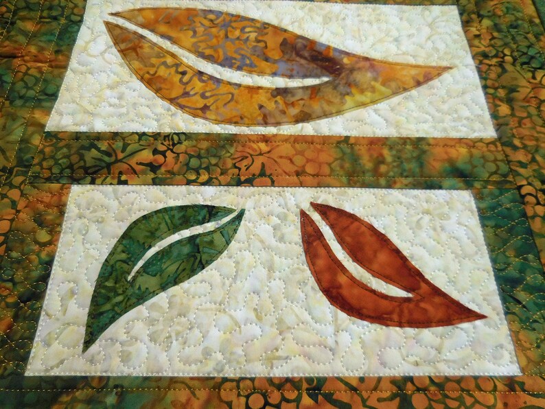 以下が含まれることがあります： 緑、オレンジ、茶色の葉のデザインが施された白い布のキルト。葉はアップリケされ、装飾的なステッチでキルティングされています。