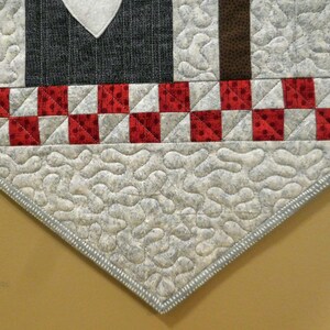 May include: A gray and white quilted table runner with a red and white checkered border. The runner has a scalloped edge and is decorated with a heart shape in the center.