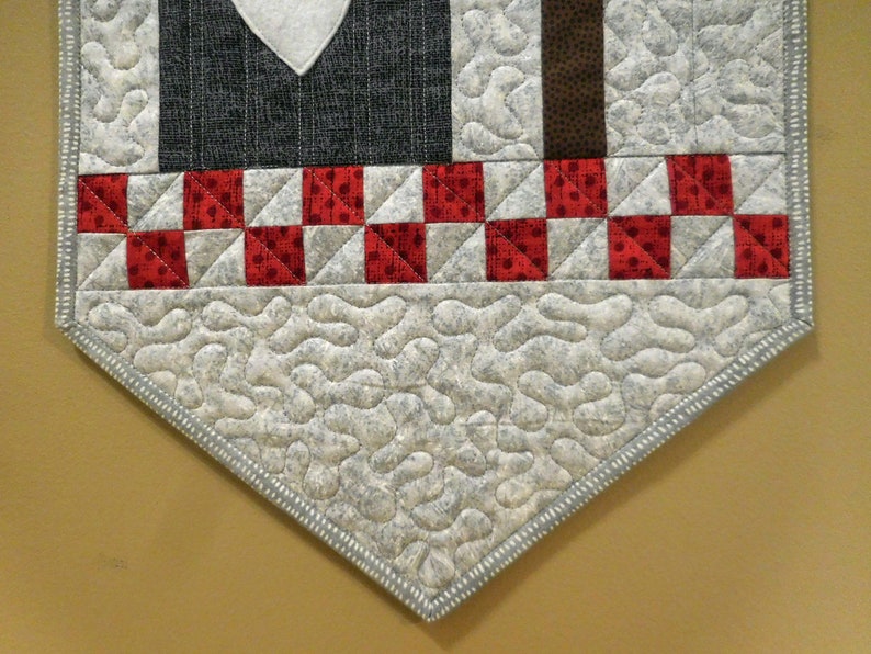 May include: A gray and white quilted table runner with a red and white checkered border. The runner has a scalloped edge and is decorated with a heart shape in the center.