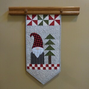 May include: A quilted wall hanging featuring a gnome with a red and black plaid hat, a white beard, and a gray body. The gnome is standing in front of a green tree with a brown trunk. The hanging is decorated with red and white checkered fabric and a white background.