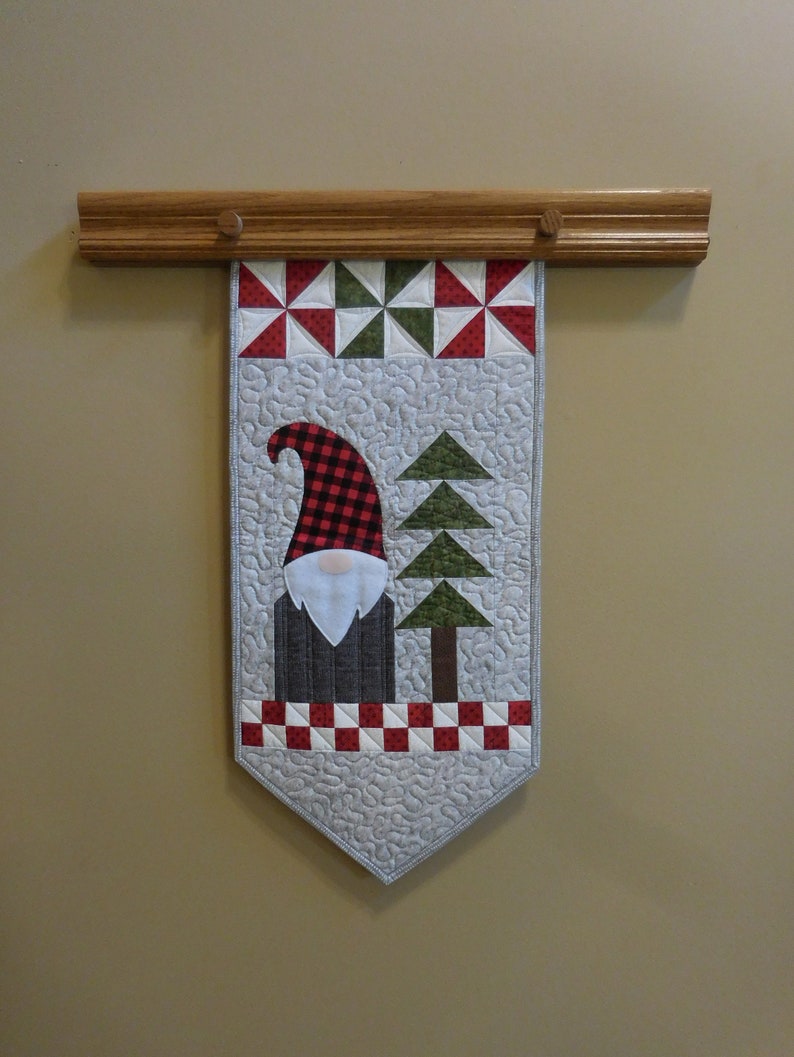 May include: A quilted wall hanging featuring a gnome with a red and black plaid hat, a white beard, and a gray body. The gnome is standing in front of a green tree with a brown trunk. The hanging is decorated with red and white checkered fabric and a white background.