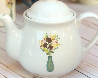 May include: White ceramic teapot with a floral design. The teapot has a lid and a spout. The floral design features a bouquet of brown, yellow, and green flowers in a blue vase.
