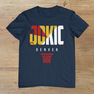 May include: A navy blue t-shirt with the word "JOKIC" in white letters with red and yellow accents. The word "DENVER" is printed below the name. A basketball hoop is printed below the text.