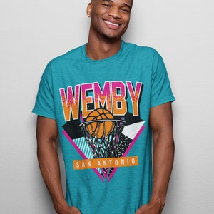 May include: A turquoise blue t-shirt with a retro-style graphic of a basketball going through a hoop. The graphic includes the text "WEMBY" and "SAN ANTONIO" in pink and white.