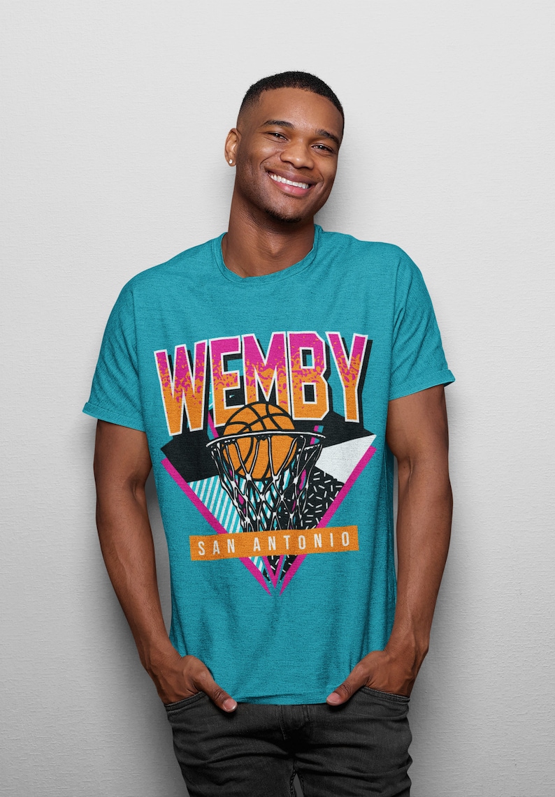 May include: A turquoise blue t-shirt with a retro-style graphic of a basketball going through a hoop. The graphic includes the text "WEMBY" and "SAN ANTONIO" in pink and white.