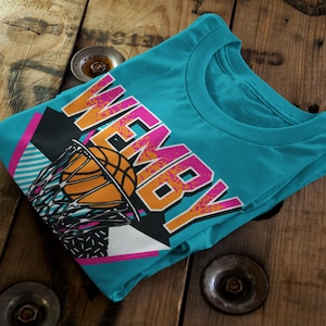 May include: A turquoise blue t-shirt with a retro-style graphic of a basketball going through a hoop. The graphic includes the word "WEMBY" in pink and orange letters.