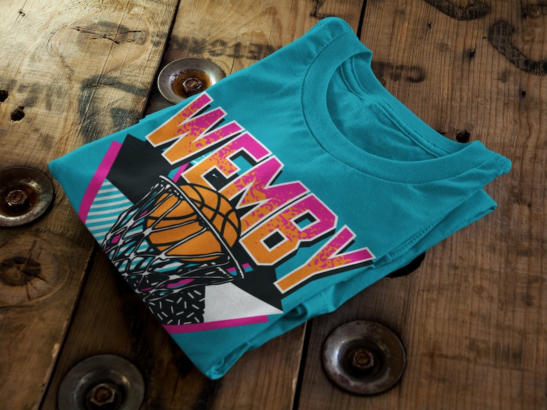May include: A turquoise blue t-shirt with a retro-style graphic of a basketball going through a hoop. The graphic includes the word "WEMBY" in pink and orange letters.
