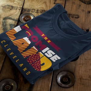 May include: A navy blue t-shirt with a graphic design featuring the words "The Promise Land" in white and yellow with a red and yellow geometric pattern. The text "Cleveland" is printed in white below the graphic.