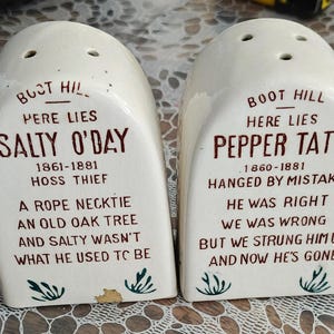 May include: A pair of vintage ceramic salt and pepper shakers. Each shaker is off-white with a rounded top and three holes. The text on the shakers reads "Boot Hill, Here Lies" followed by names and epitaphs in brown lettering. Decorative sprigs are at the bottom.