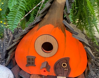 May include: A whimsical, handmade birdhouse shaped like a pumpkin. The birdhouse features an orange body, a circular entry hole, and a wooden shingle roof. Decorative elements include a small window and a door with a keyhole. A string is attached for hanging.