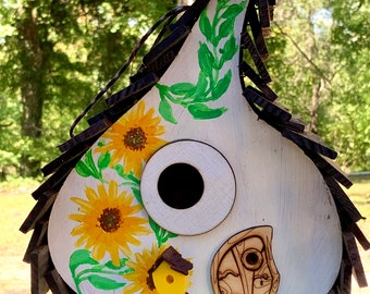 May include: A white birdhouse shaped like a teardrop with a black roof and a round entrance hole. The birdhouse is decorated with painted sunflowers, green leaves, and a small wooden door with a whimsical design.