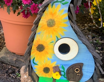 May include: A blue birdhouse shaped like a teardrop with a sunflower design. The birdhouse has a small door with a keyhole and a round opening for birds to enter. The birdhouse is sitting on a rock.