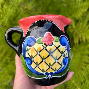 Puede incluir: Jarra de cerámica negra con borde rojo y un diseño floral colorido. El diseño presenta un patrón de cuadros amarillos y blancos con detalles azules y verdes.