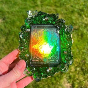 May include: A green resin art piece encasing a 'Forest' trading card. The card features a colorful, abstract tree design. The resin has a crystalline texture and is held by a hand against a blurred green background.