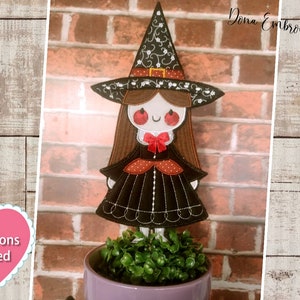 May include: A black and red witch doll with a black hat and a red bow. The doll is standing in a purple pot with green plants.
