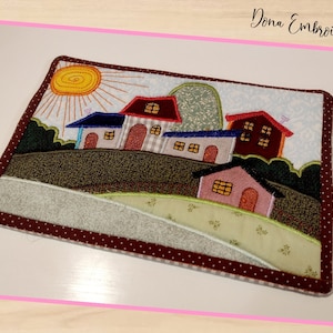 Pode incluir: Uma tapeçaria bordada colorida que apresenta uma cena de vila caprichosa com casas, árvores e um sol. As casas são vermelhas, azuis e cor-de-rosa com janelas e portas brancas. As árvores são verdes e castanhas. O sol é amarelo com raios laranja. O fundo é um céu azul claro com nuvens brancas. A tapeçaria é emoldurada com uma borda castanha com bolinhas brancas.