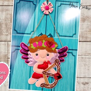 May include: A pink and purple felt angel with brown hair, wearing a red dress and holding a bow and arrow. The angel is hanging from a string on a blue door. The text "Instructions Included" is in a pink heart shape.