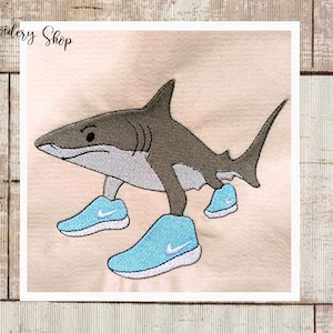 Pode incluir: Imagem bordada de um tubarão usando tênis azul claro com detalhes brancos. O tubarão é cinza e branco, com contornos pretos. A imagem está em um fundo bege claro. O texto "Dona Embroidery Shop" está no canto superior esquerdo.