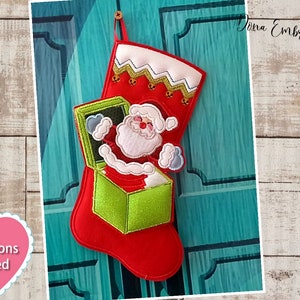 以下が含まれることがあります： 緑色の箱と、上から顔を出しているサンタクロースが付いた赤いクリスマスストッキング。ストッキングには白いジグザグの縁取りと金のベルが付いています。