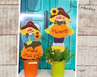 May include: Two scarecrow figures holding pumpkins with embroidered text. One pumpkin says "Welcome" and the other says "Happy Fall Y'all". The figures are in orange, brown, and blue fabric and are placed in orange and green buckets.