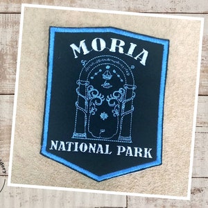 May include: A black and blue embroidered patch with the text "Moria National Park" and a white outline of a gate with a crown on top.