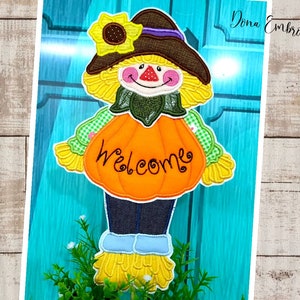 May include: A colorful embroidered scarecrow holding a pumpkin with the word "welcome" on it. The scarecrow is wearing a brown hat, a green shirt, and blue pants. The pumpkin is orange with a green stem.