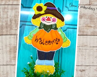 May include: A colorful embroidered scarecrow holding a pumpkin with the word "welcome" on it. The scarecrow is wearing a brown hat, a green shirt, and blue pants. The pumpkin is orange with a green stem.