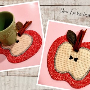 May include: A red apple-shaped fabric coaster with a brown cinnamon stick and green leaf on top. The coaster has a white center with a small black heart design. The coaster is on a pink surface.