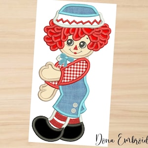 May include: Embroidered design of a red-haired rag doll with a blue and white hat, red and white checkered shirt, blue overalls, and black shoes. The doll is smiling and has a blue bow on its chest.