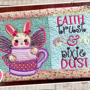 May include: A quilted wall hanging with a pink and green bunny holding a stack of purple cups. The text "Faith Trust & Pixie Dust" is embroidered in pink and black on a light blue background.