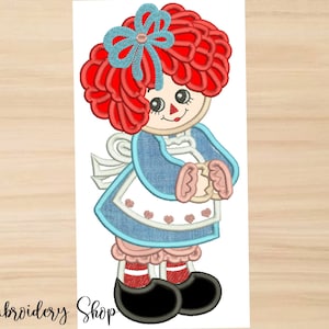 Puede incluir: Diseño bordado de una muñeca de trapo de pelo rojo con un vestido azul, un delantal blanco y zapatos negros. La muñeca tiene un lazo azul en el pelo y tiene las manos juntas.